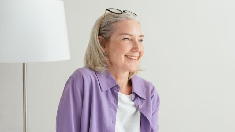 Smiling older woman with gray hair wearing a white shirt and lavender button-down, glasses resting on her head, standing near a floor lamp against a light-colored wall, appearing relaxed and content in a bright indoor setting
