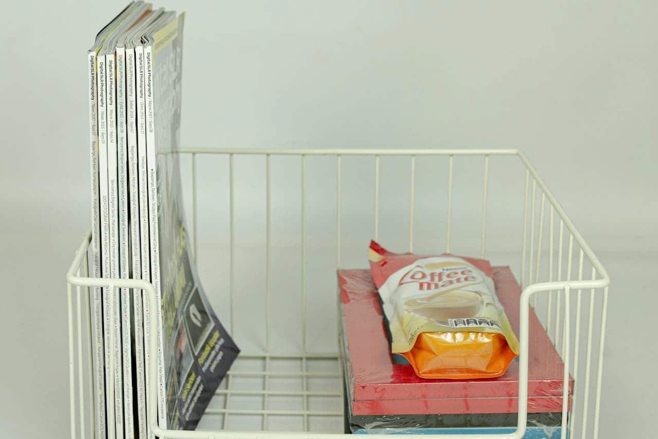 White wire basket containing several magazines or thin books standing upright beside Coffee-mate creamer on pink paper