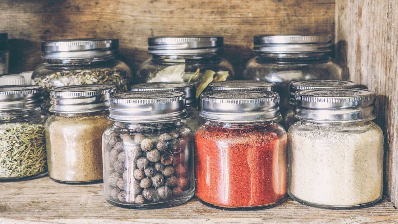 A rustic wooden shelf holding small glass jars filled with various colorful spices and herbs, each sealed with a metal lid