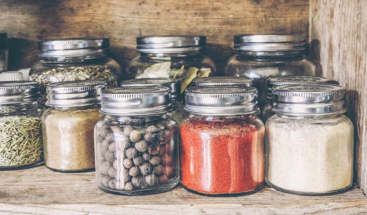 A rustic wooden shelf holding small glass jars filled with various colorful spices and herbs, each sealed with a metal lid