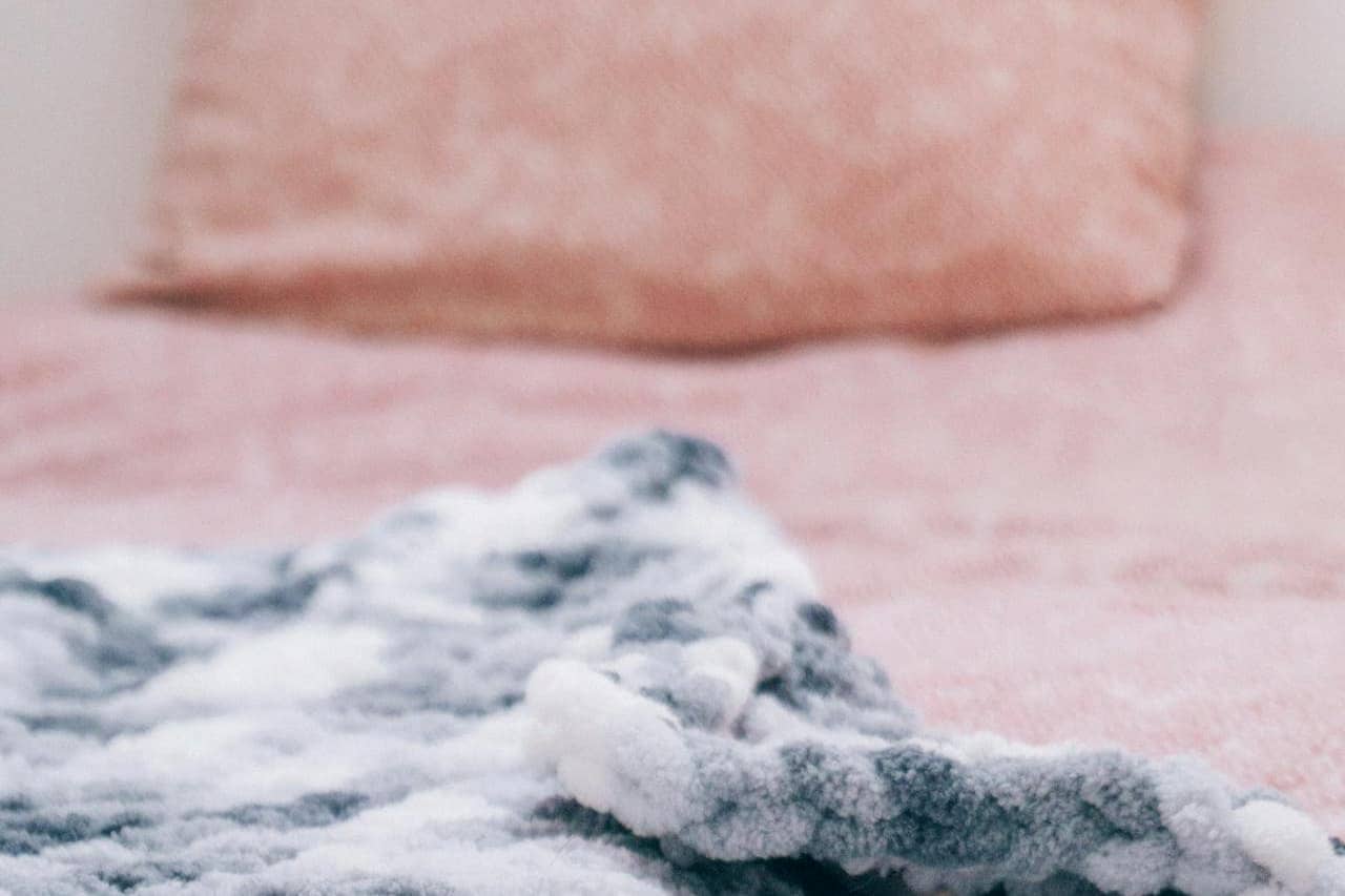 Chunky knit wool blanket with white pom-poms resting on pink bedding, creating cozy bedroom contrast