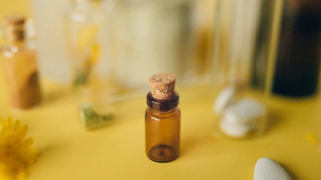 Small amber glass bottle with cork stopper, placed on a yellow surface, surrounded by white stones, yellow flowers, and blurred glassware in the background, soft natural lighting, minimalistic and warm aesthetic