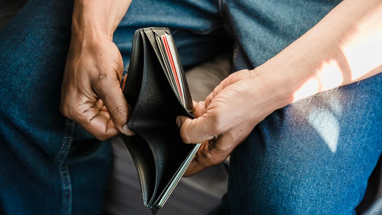 Close-up of a person holding an empty black wallet open, revealing no cash inside, while sitting on a bed in blue jeans