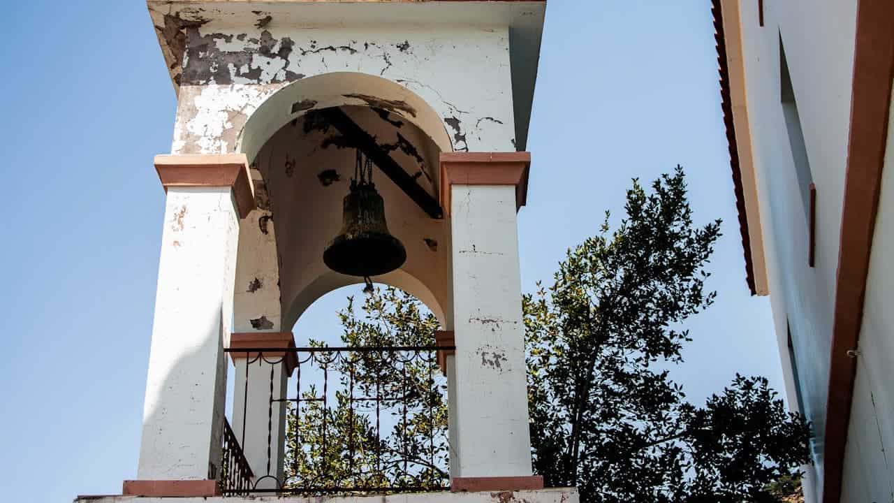 Weathered church bell tower with severely peeling white paint showing deterioration, featuring hanging bell and surrounding green trees