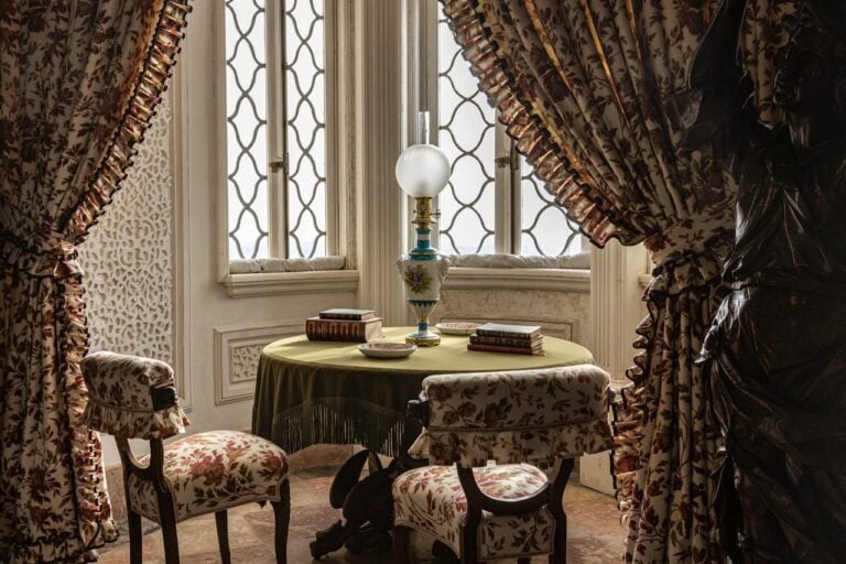 Vintage room with floral curtains, upholstered chairs, round table, oil lamp, antique books, and ornate windows with patterned glass