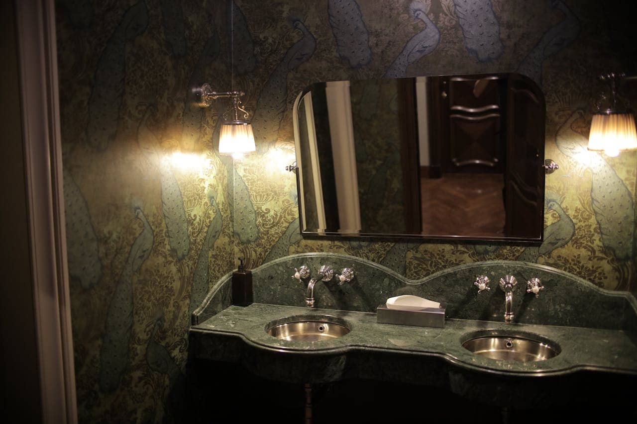 Elegant bathroom with green marble double vanity, ornate wallpaper, sconce lighting, and rectangular mirror above twin sinks