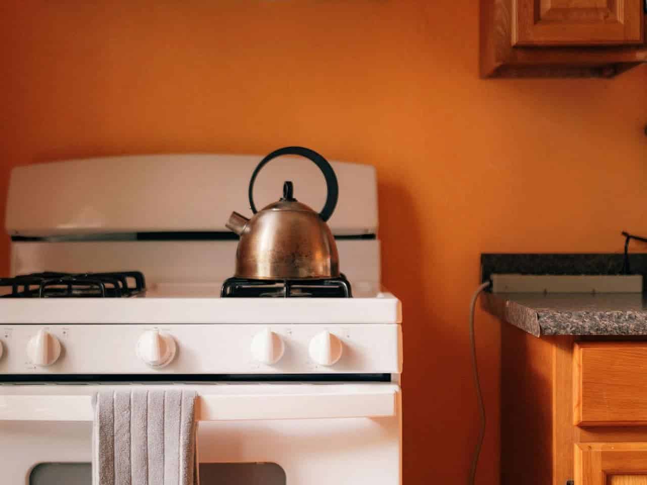 A white gas stove with a kettle on top, a grey towel hanging from the stove, wooden cabinets on the right, an orange wall in the background, clean and minimalist kitchen setting