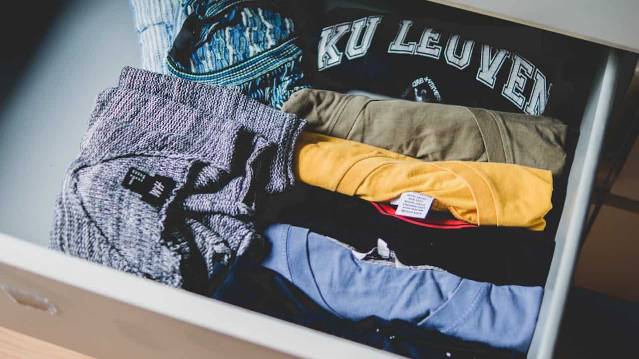 Open drawer containing folded clothing items including a "KU LEUVEN" sweatshirt, gray sweater, yellow shirt, and blue fabric