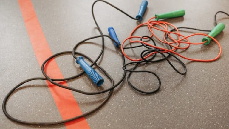 Several colorful jump ropes with green, blue, and red handles tangled on a gym floor, near a red line marking, on a textured rubber surface