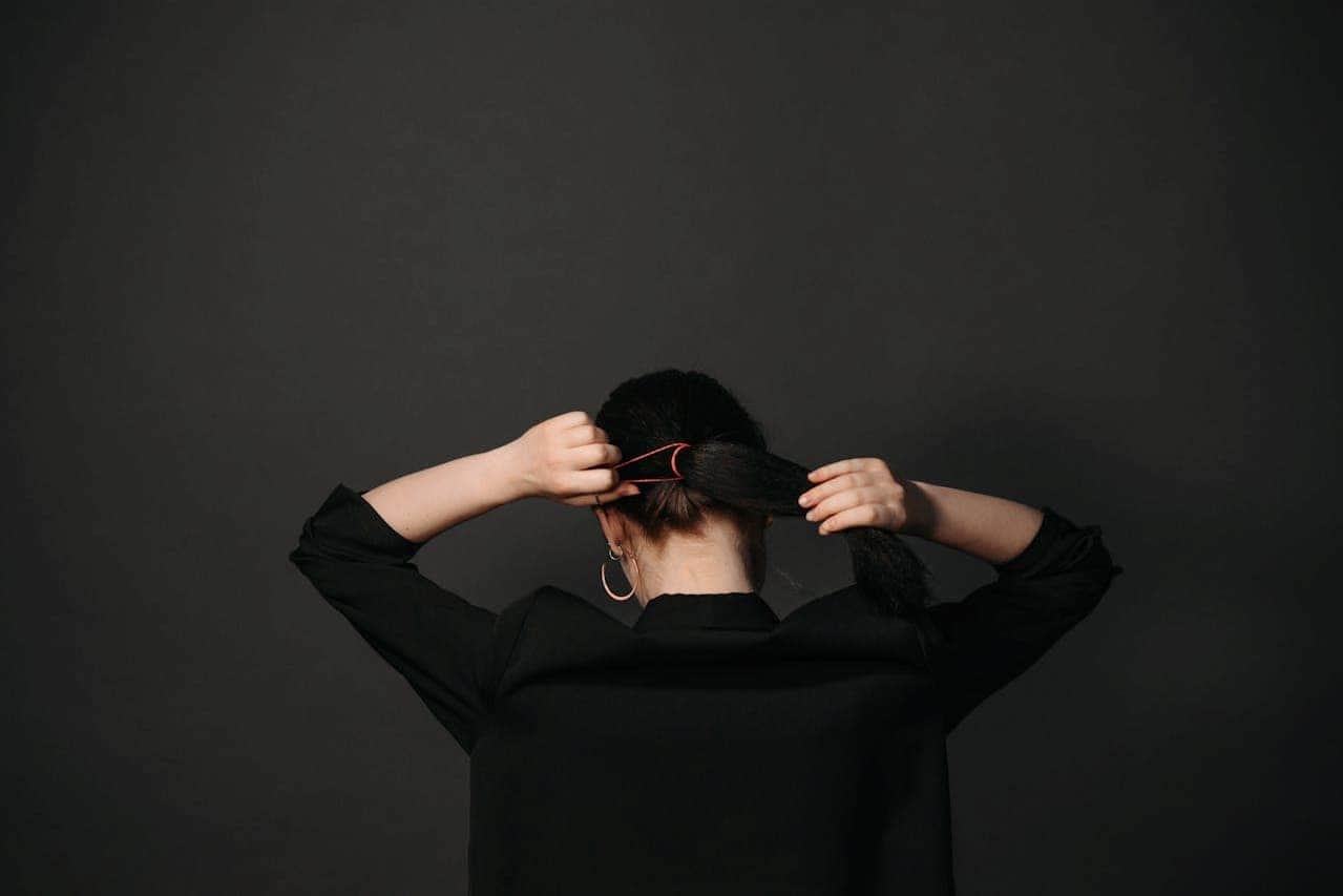 Back view of person with dark hair in black clothing tying hair with red hair tie against dark background