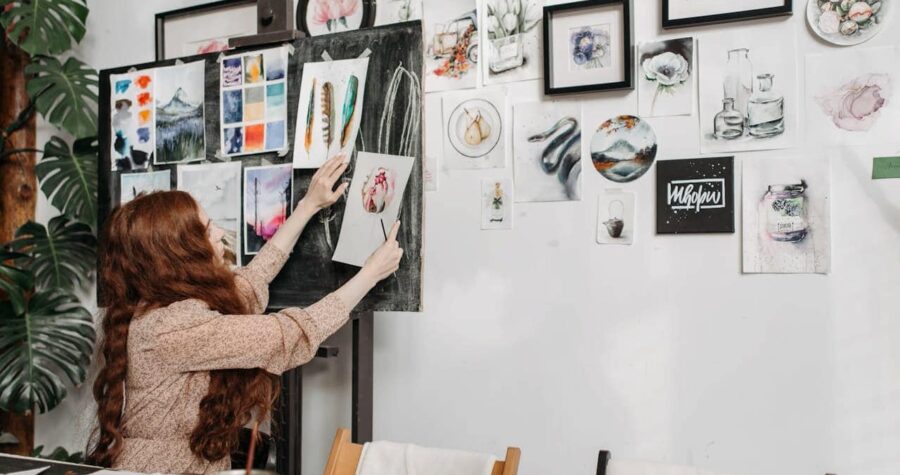 Woman with long red hair arranging artwork on white gallery wall filled with various paintings, sketches, and framed pieces
