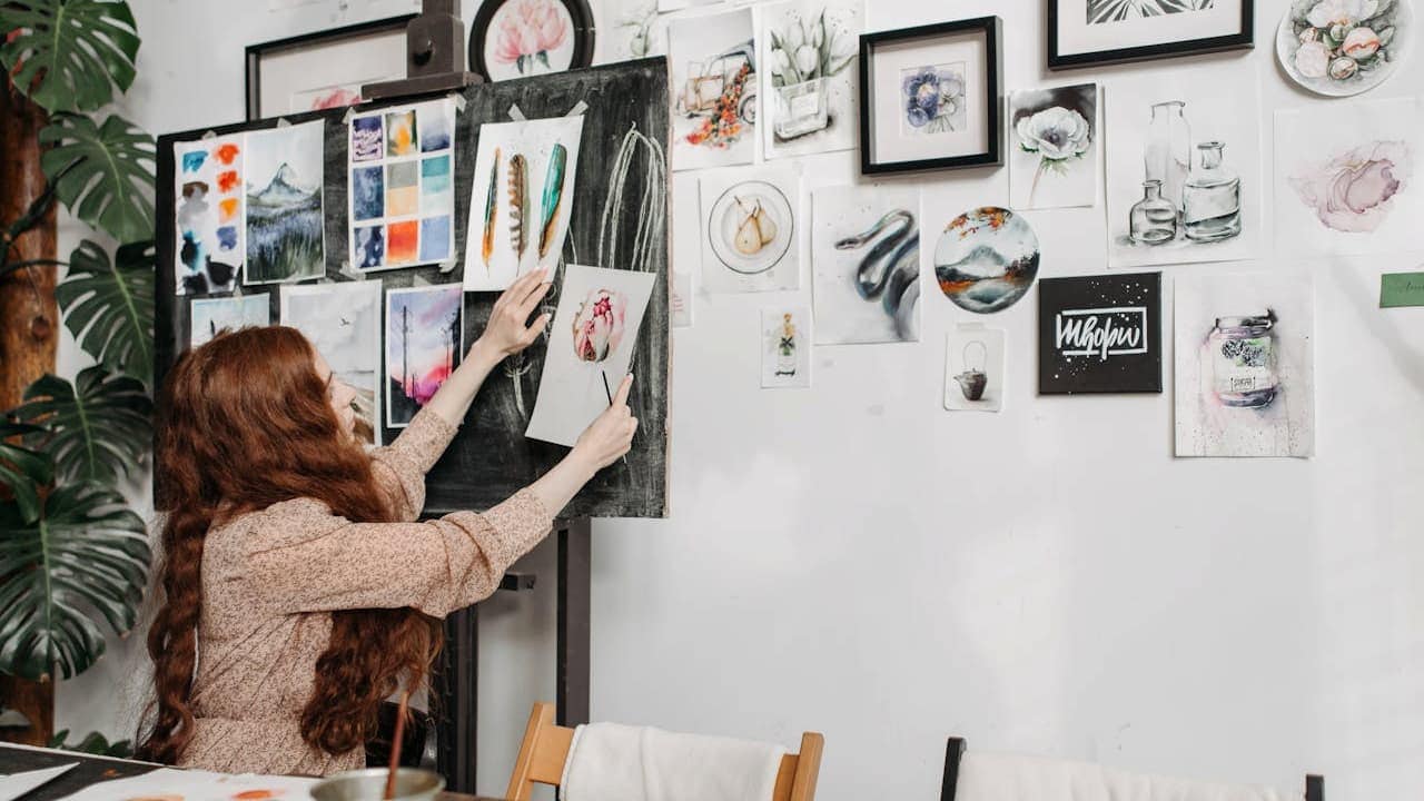 Woman with long red hair arranging artwork on white gallery wall filled with various paintings, sketches, and framed pieces