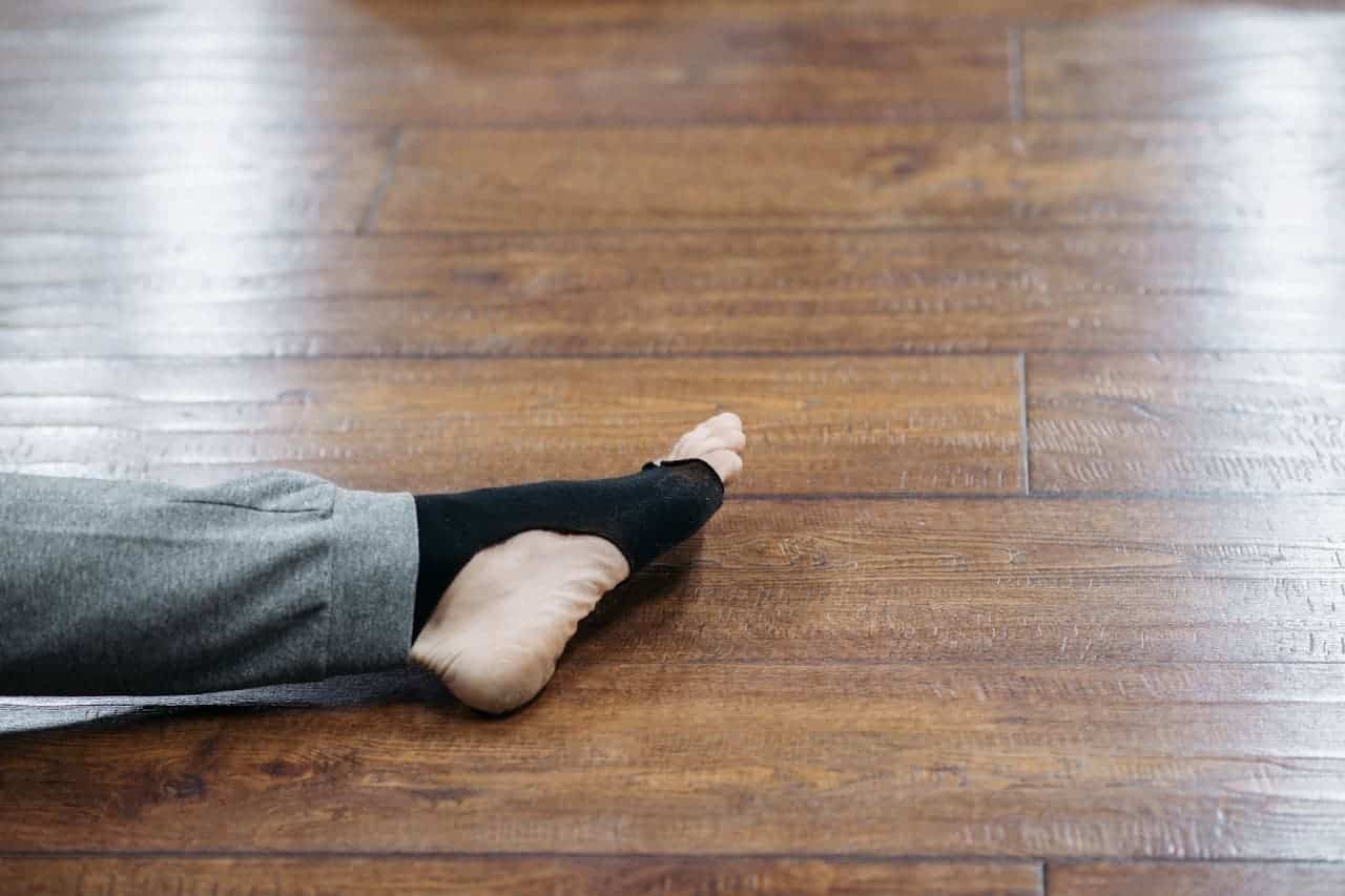 Person's foot with black holey sock resting on polished wooden floor, gray pant leg visible