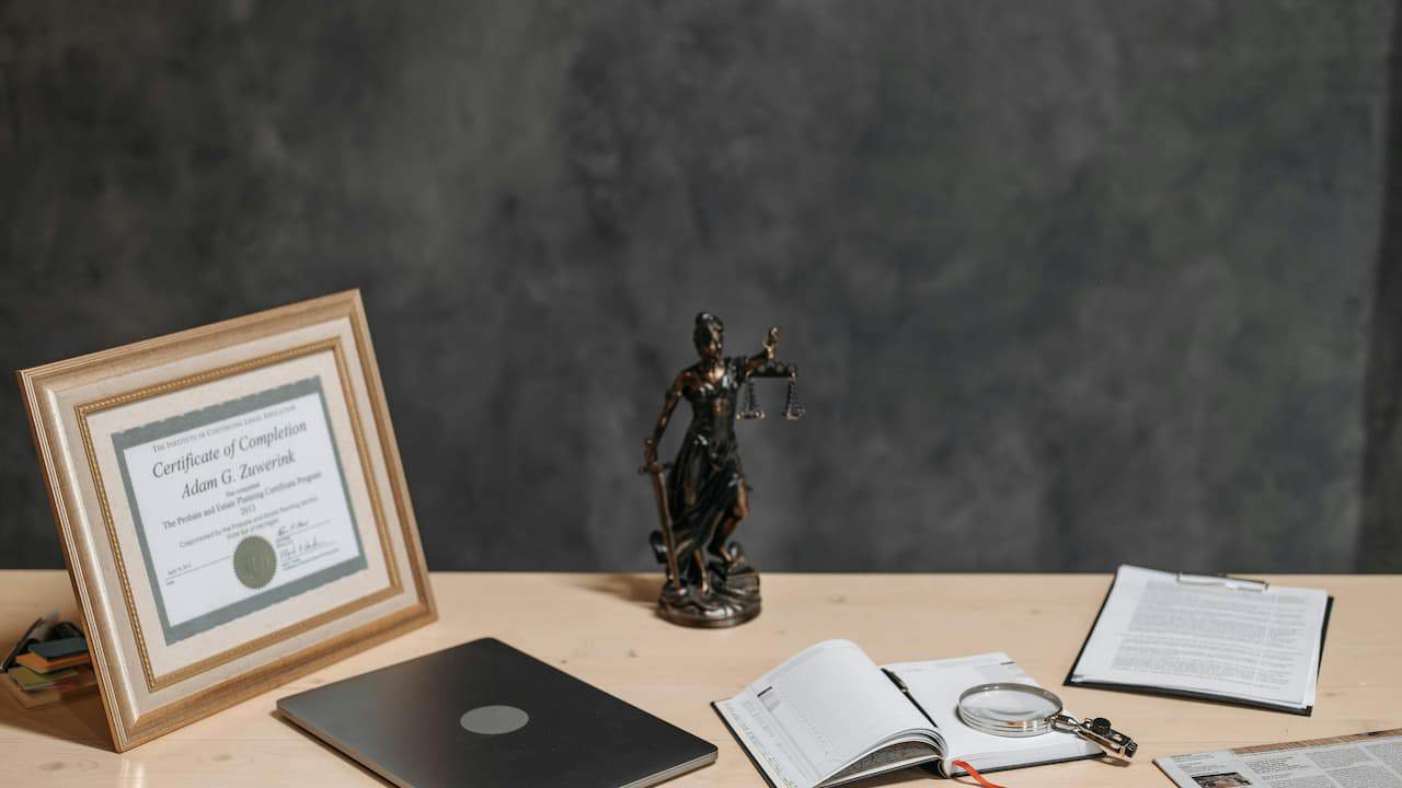 A framed award certificate displayed on a table alongside a black abstract sculpture, an open book, and a clipped stack of papers, all arranged neatly on a wooden surface