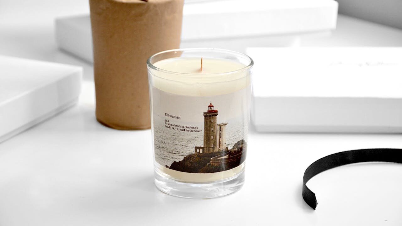 Scented soy candle with lighthouse label on white desk, surrounded by wrapped package, ribbon, and white boxes in soft lighting