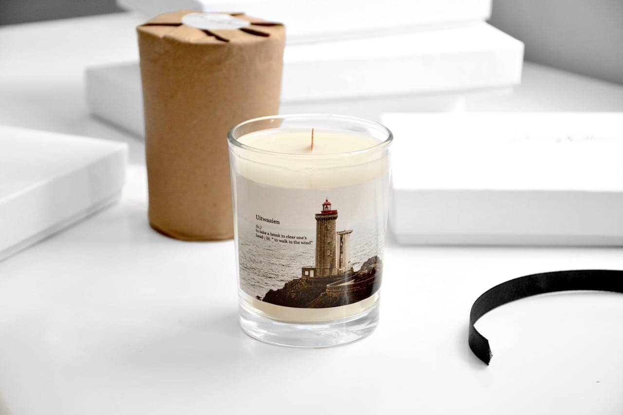 Scented soy candle with lighthouse label on white desk, surrounded by wrapped package, ribbon, and white boxes in soft lighting