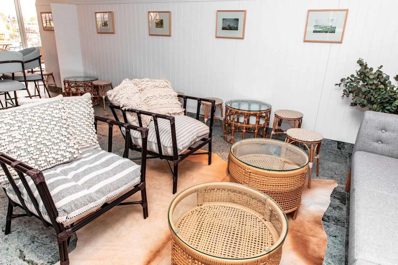 Bright sunroom with rattan furniture, glass-top tables, patterned cushions, white paneled walls, framed artwork, and potted plants