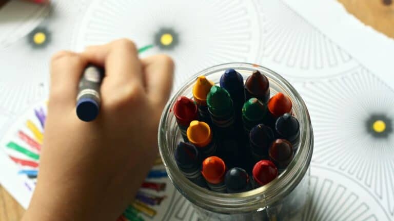 A child’s hand reaching into a container filled with colorful crayons, actively coloring on a drawing with bright, varied hues, close-up with natural light