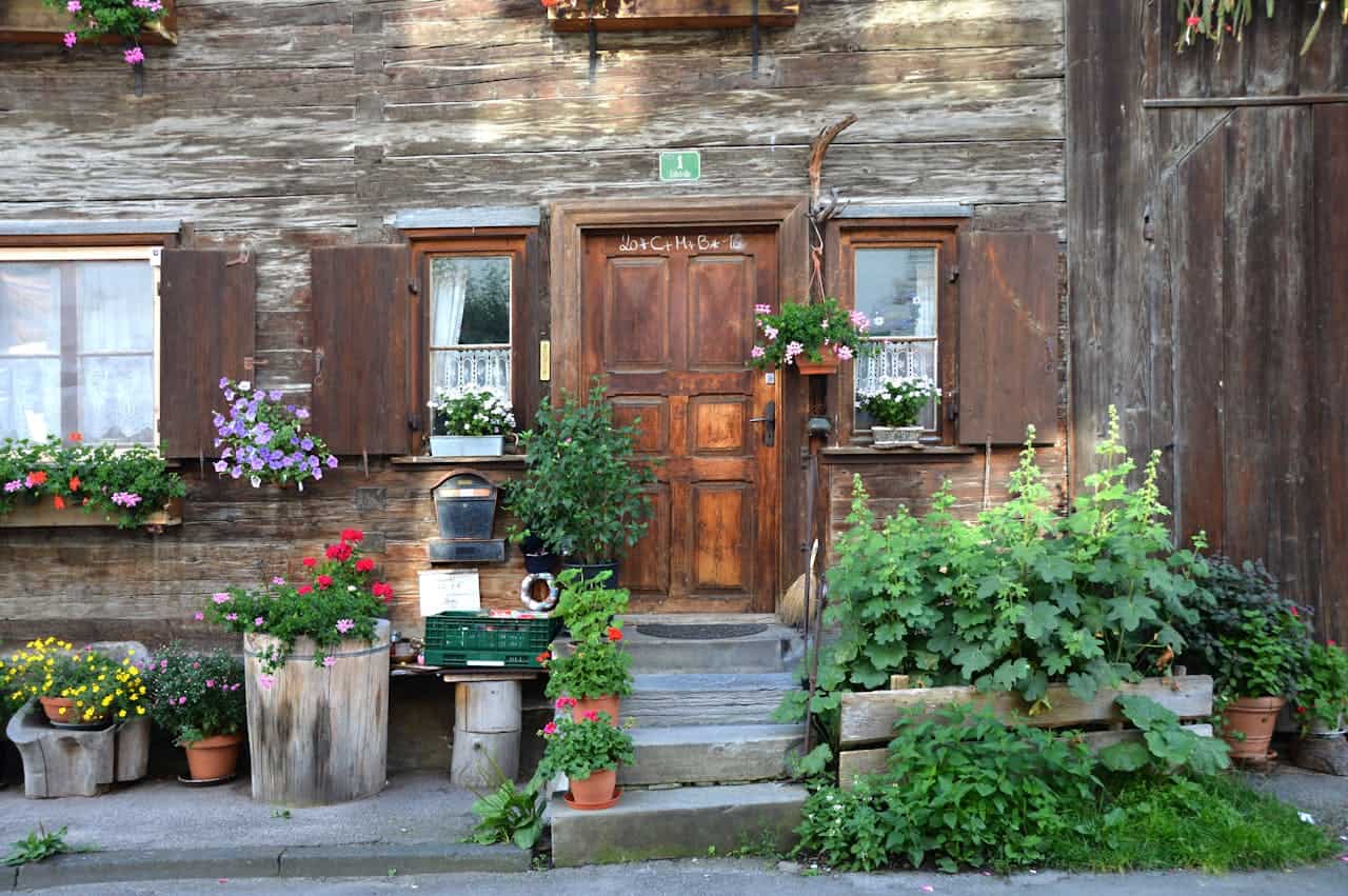 Rustic wooden cabin entrance with wooden shutters, colorful flower boxes, potted plants including perennials , and weathered steps leading to a brown door