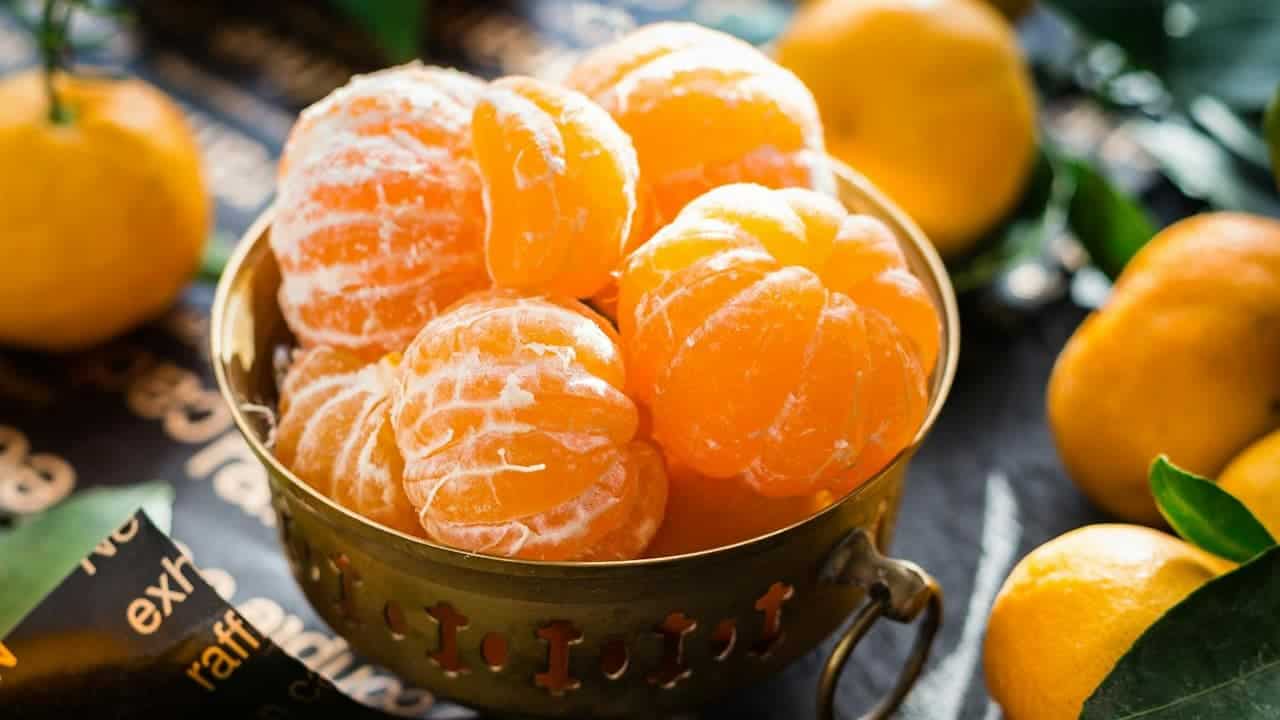 Clementines in a brass bowl with whole unpeeled clementines around, lit by bright natural light on a patterned surface