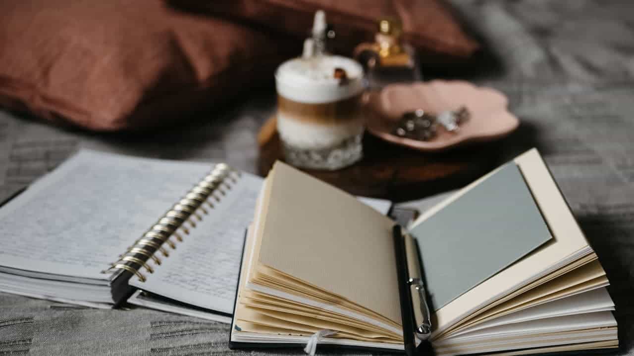 : Open notebook and book on wooden table with layered coffee drink, copper tray with treats, and cozy blanket nearby