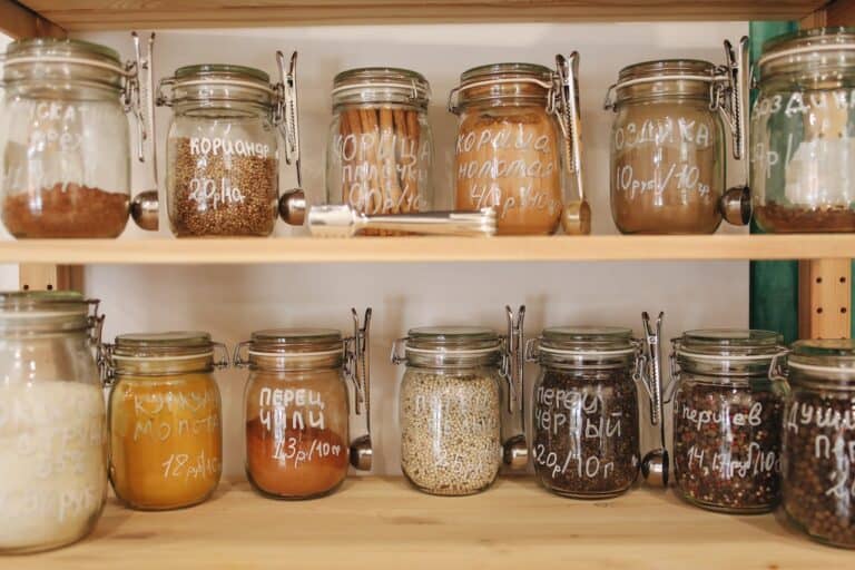 Glass jars filled with various spices and grains labeled in Russian, arranged neatly on wooden kitchen shelves