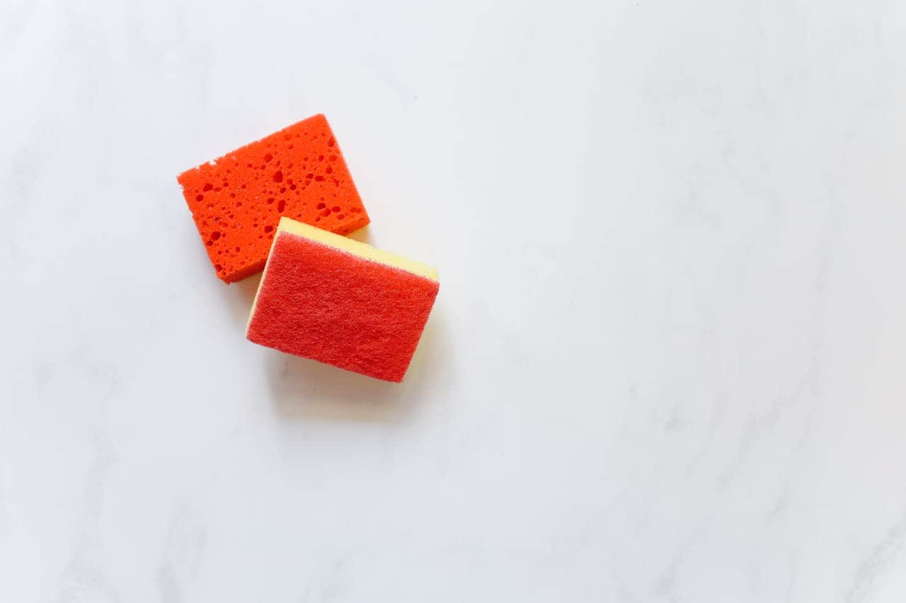 Two orange kitchen sponges on white marble surface - one cellular foam type, one with scrubbing pad attached