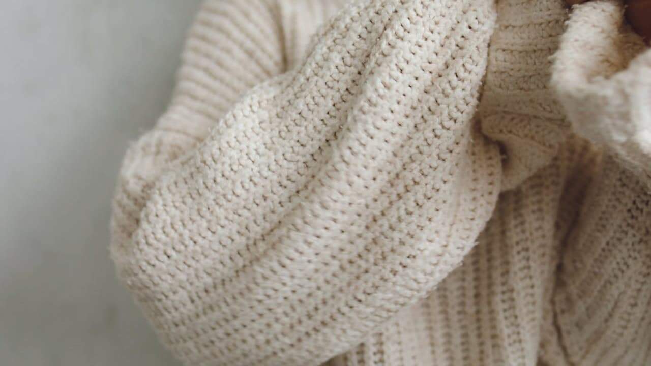 Close-up of cream-colored knitted sweater showing detailed cable-knit pattern and ribbed collar texture in soft natural lighting