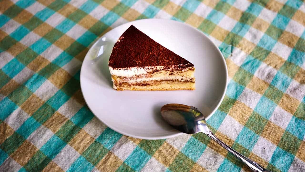 Slice of tiramisu cake on white plate with spoon, served on turquoise and yellow checkered tablecloth