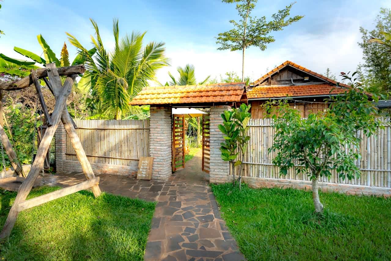 Tropical garden entrance with stone pathway leading to brick gateway, bamboo edging and tiled-roof cottage surrounded by palm trees