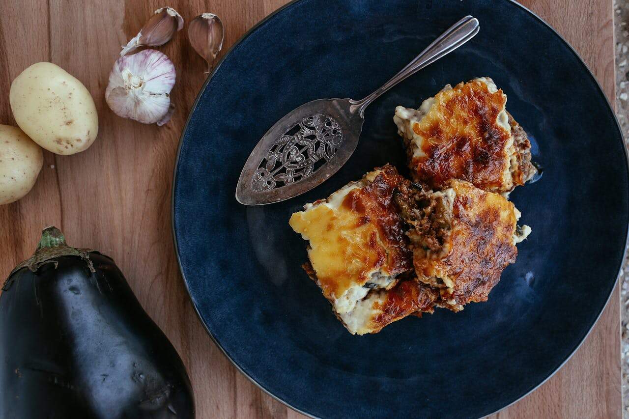 Navy blue plate with baked casserole serving and decorative spoon on wooden cutting board beside eggplant, potatoes, and garlic