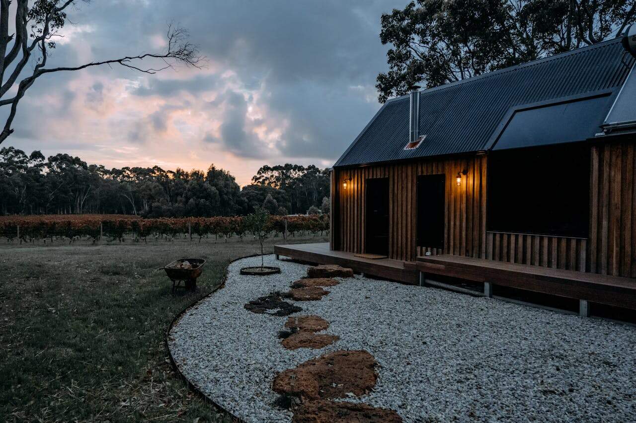 Wooden cabin with metal roof beside gravel garden path with flexible steel edging, overlooking vineyard at sunset with dramatic cloudy sky