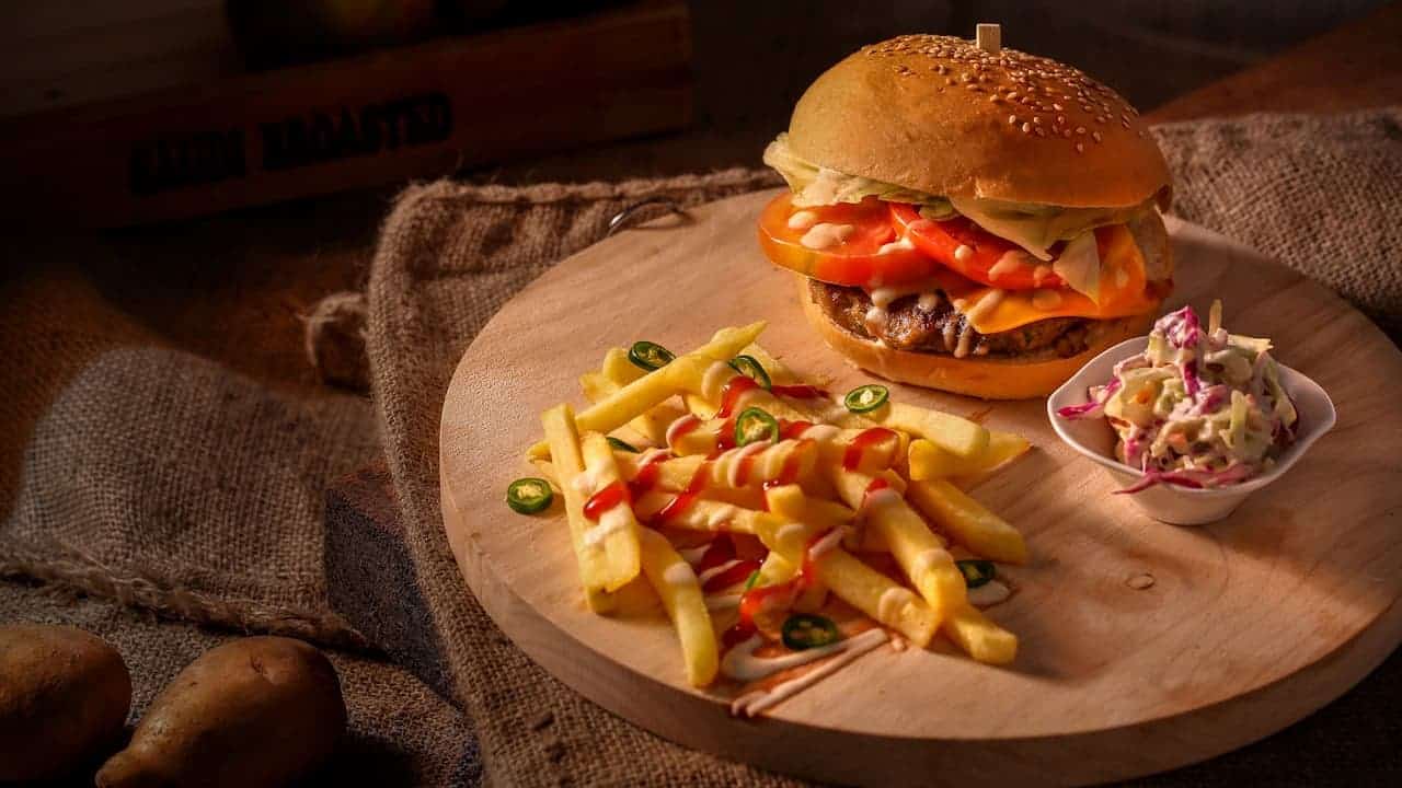 Gourmet burger with sesame seed bun, loaded fries with toppings, and coleslaw side dish served on circular wooden cheese boards