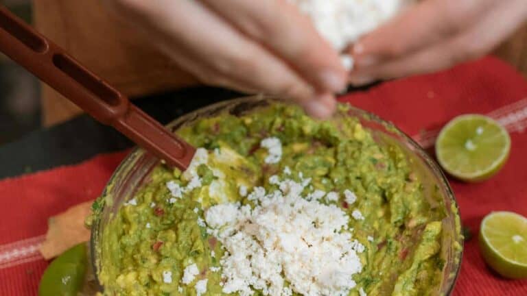 Person crumbling white cheese over a bowl of guacamole, with sliced limes, avocado, and chili peppers