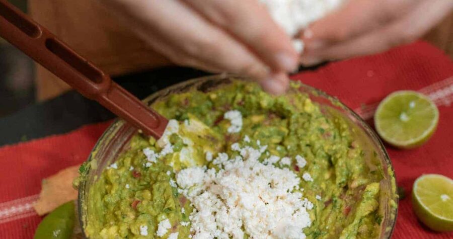 Person crumbling white cheese over a bowl of guacamole, with sliced limes, avocado, and chili peppers