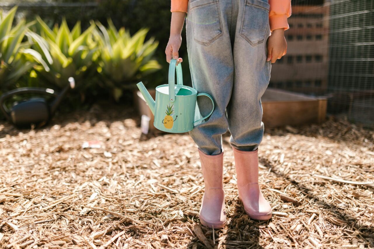 Person in rolled-up jeans and pink boots standing on mulch, holding a mint-green watering can with a pineapple sticker, garden plants in the background