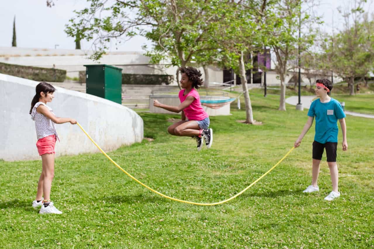 Children playing jump rope in a park. Two kids hold rope while girl in pink shirt jumps in grassy area