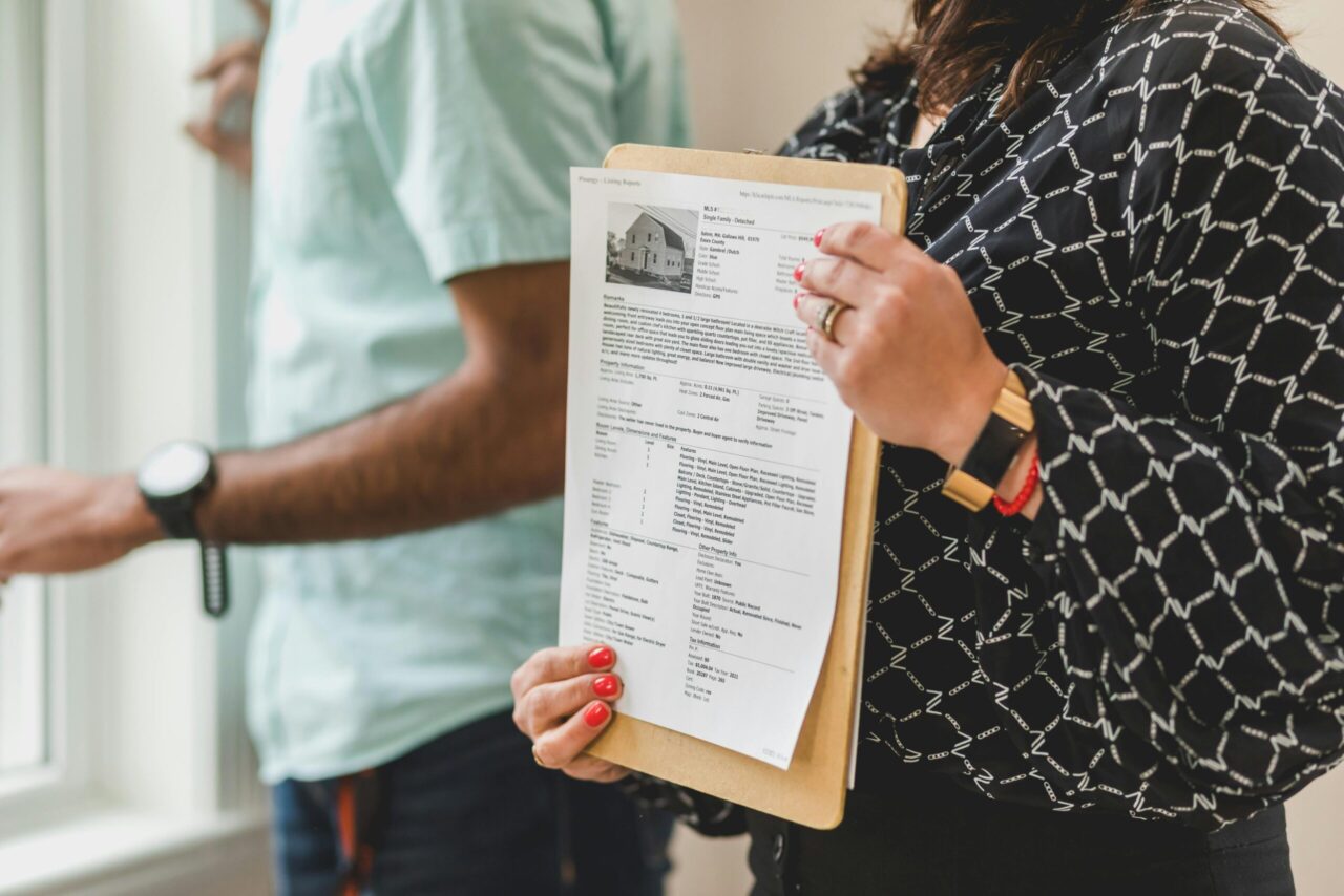 Real estate agent holding a clipboard with a printed property listing, close-up of document showing house details and photo, person in patterned black blouse, standing next to a man by a window, indoor setting during property showing