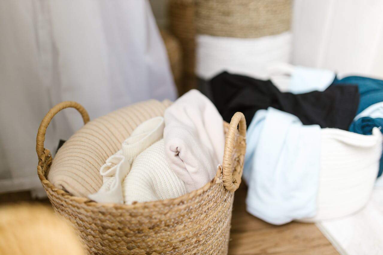Woven basket with folded cream sweaters and white clothing; unmade bed visible in background with black and blue bedding