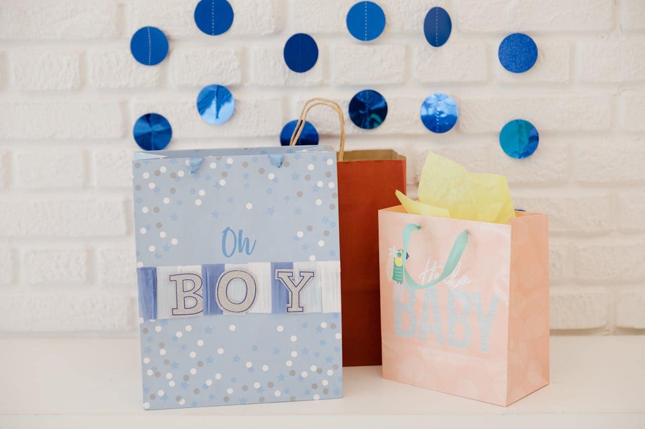 Baby shower gift bags with "Oh BOY" and "BABY" text against white brick wall decorated with blue circular garland