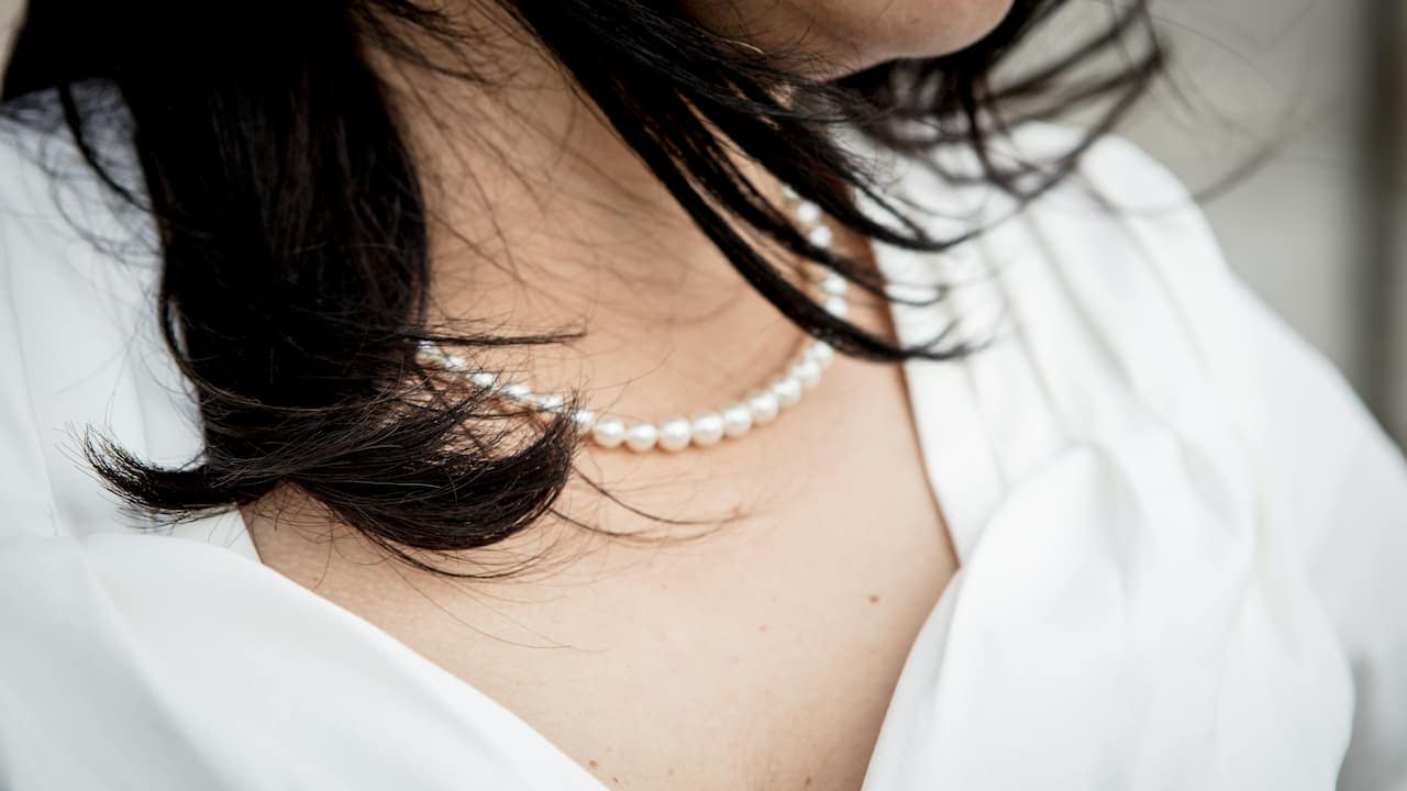 A white woman with dark hair wearing a white top and a pearl necklace, photographed against a neutral background, facing forward with a gentle expression