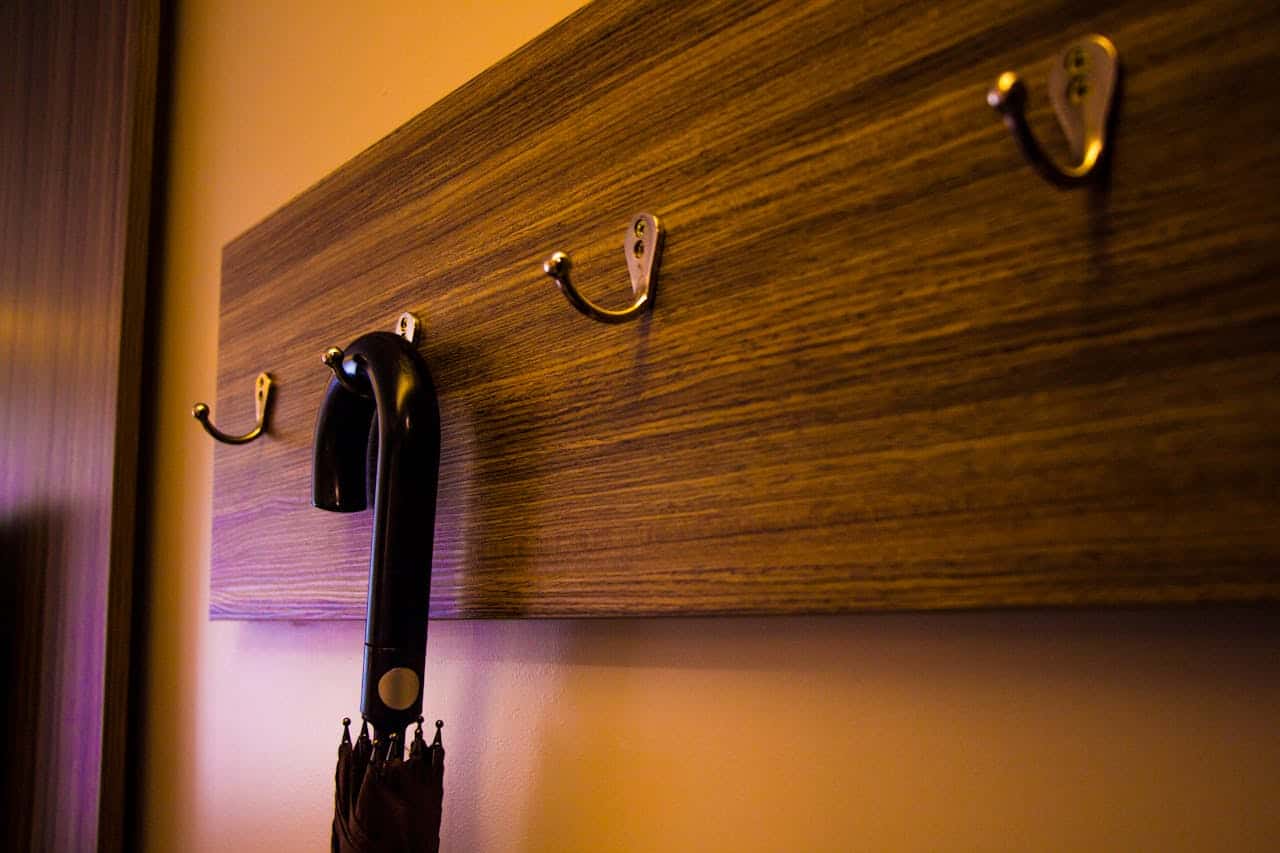 Wooden wall-mounted coat rack with four brass hooks, one holding a black umbrella, against warm-toned wall with ambient lighting