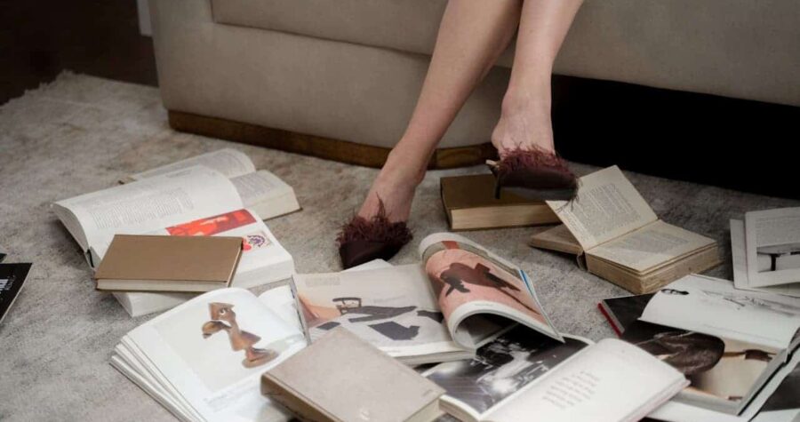 Person wearing burgundy fuzzy slippers standing among scattered open books and magazines on light carpet near beige couch
