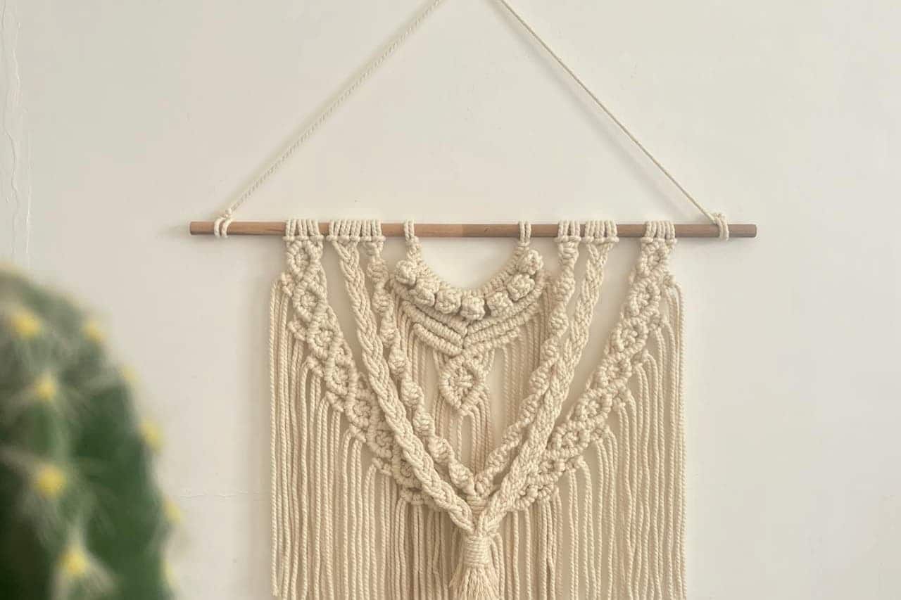 Handcrafted woven wall tapestry hanging with intricate knotted design on wooden dowel against white wall, partially showing houseplant