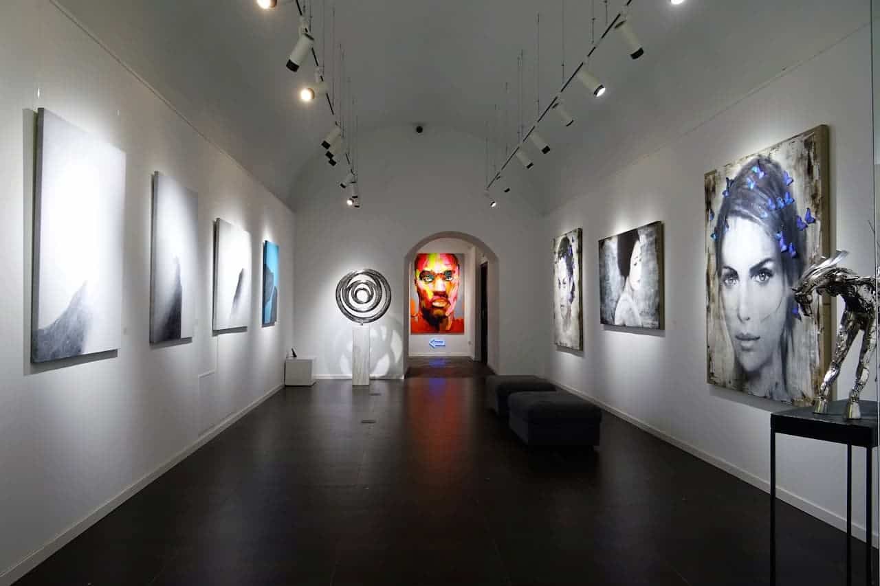 Long art gallery with white walls featuring abstract paintings, portraits, and sculptures. Track lighting illuminates artwork along both sides
