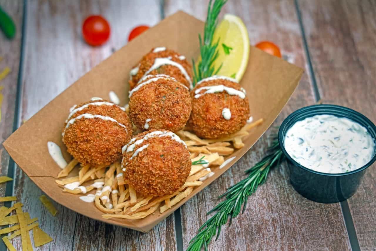Five golden-brown salmon croquettes drizzled with white sauce, served on a bed of crispy fried strips in a brown paper tray, garnished with rosemary and lemon wedge, paired with a cup of creamy dipping sauce on a rustic wooden surface