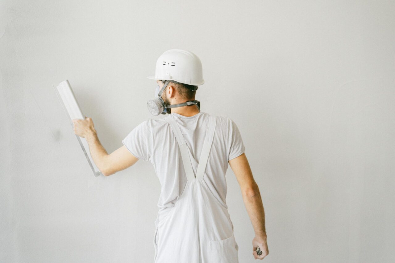 Construction worker wearing white overalls, a hard hat, and a respirator mask, smoothing a white wall with a trowel, facing away from the camera, clean and minimal indoor setting