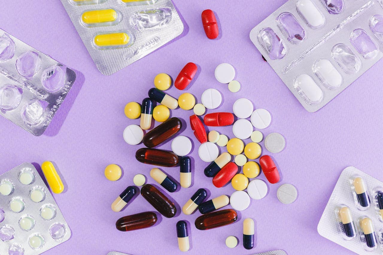 Assorted medication pills and capsules scattered on purple background, surrounded by empty blister packs of various pharmaceuticals