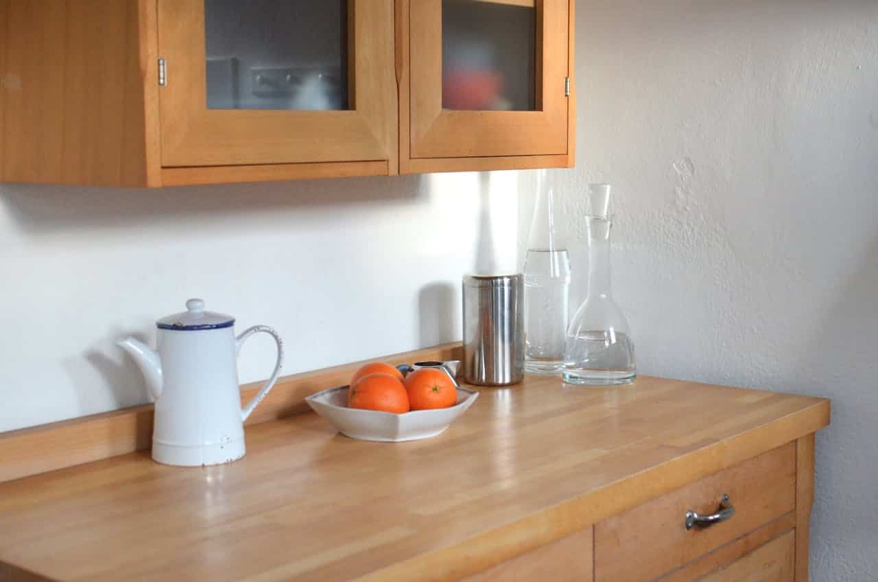 Orange wood kitchen counter with white teapot, oranges in dish, metal canister, glass decanters, and upper cabinets with glass doors