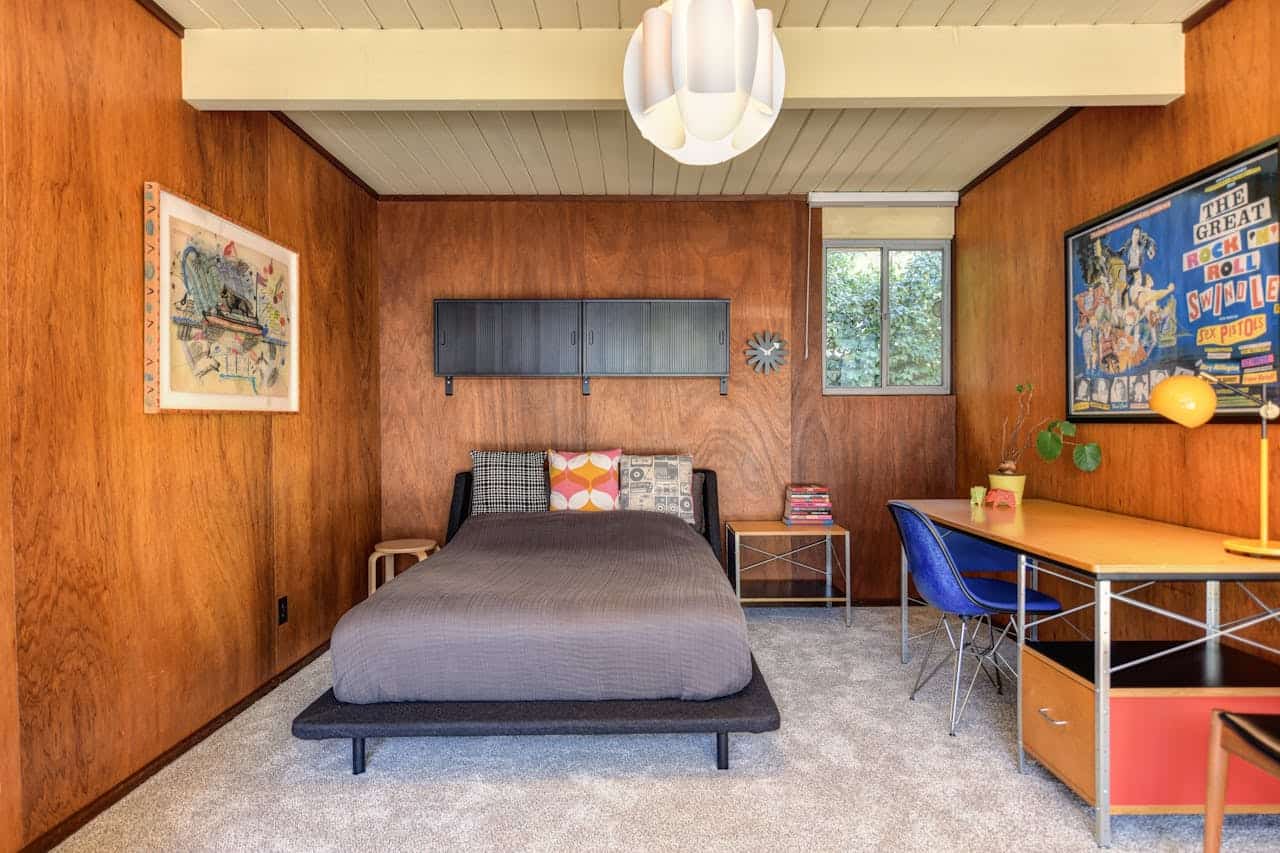 Mid-century modern bedroom with wood-paneled walls, single bed, desk with blue chair, and retro wall art